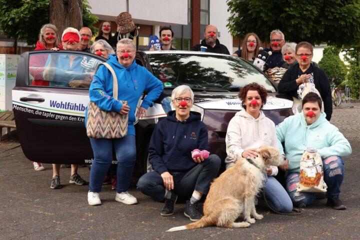 Aktivistengruppe trägt rote Clownsnasen vor einem Auto, das mit dem Slogan "Wohlfühlbote" beschriftet ist. Sie setzen sich für den Kampf gegen den Krebs ein.