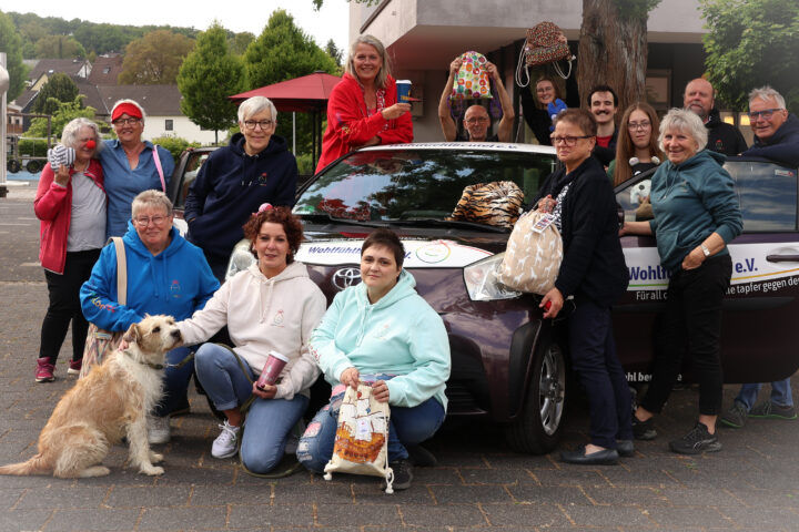 Gruppenfoto von Mitgliedern des Vereins, die sich gegen Krebs engagieren. Sie stehen vor einem Auto mit dem Vereinslogo und halten verschiedene Taschen. Ein Hund sitzt vorne.