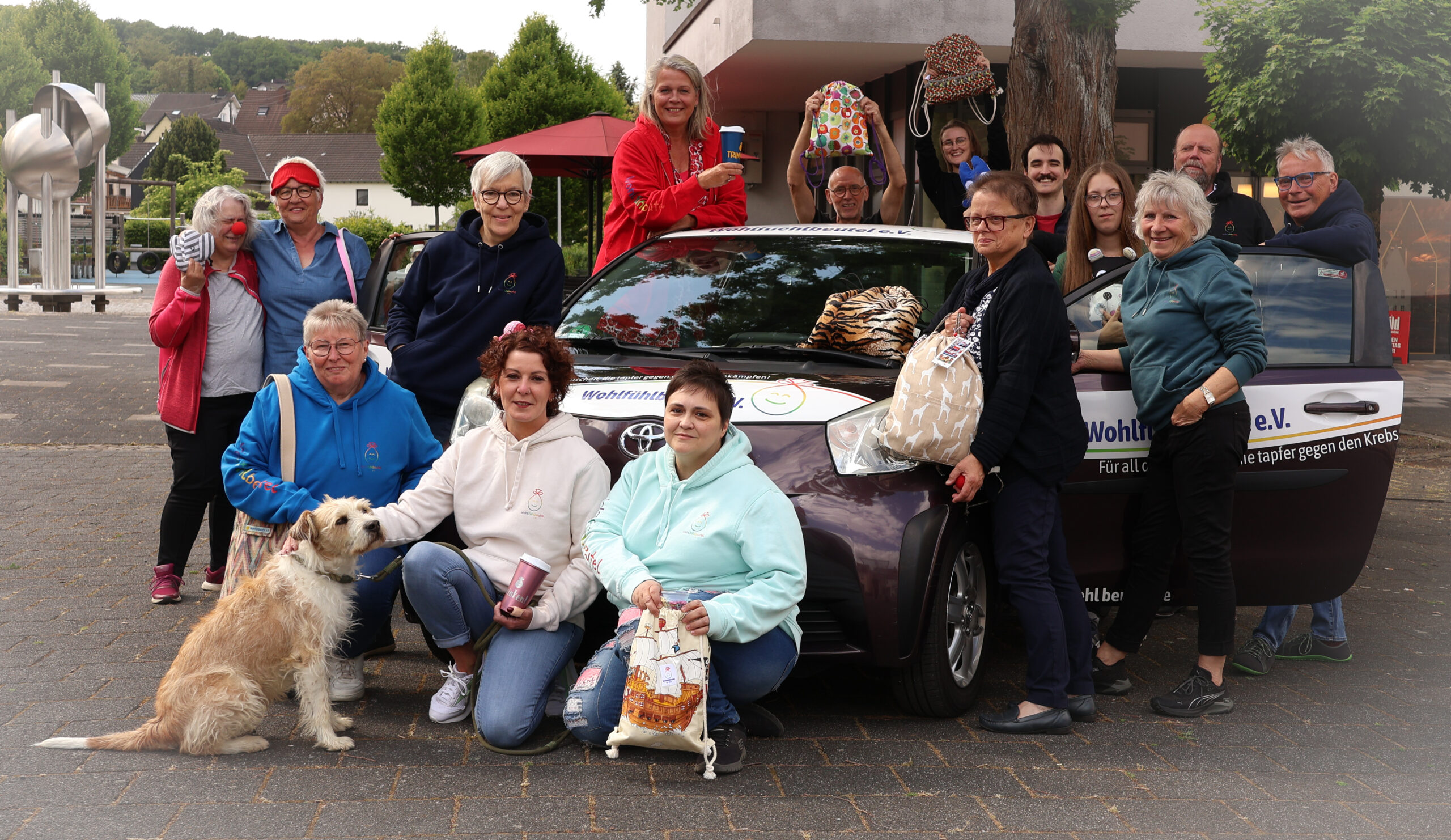 IMG_4680 (1) Gruppenfoto von Mitgliedern des Vereins, die sich gegen Krebs engagieren. Sie stehen vor einem Auto mit dem Vereinslogo und halten verschiedene Taschen. Ein Hund sitzt vorne.