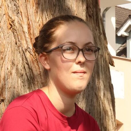Lilly Goldberg Junge Frau in rotem T-Shirt mit Hundemotiv, sitzt entspannt auf einer Bank unter einem Baum, trägt schwarze Leggings und sportliche Schuhe.