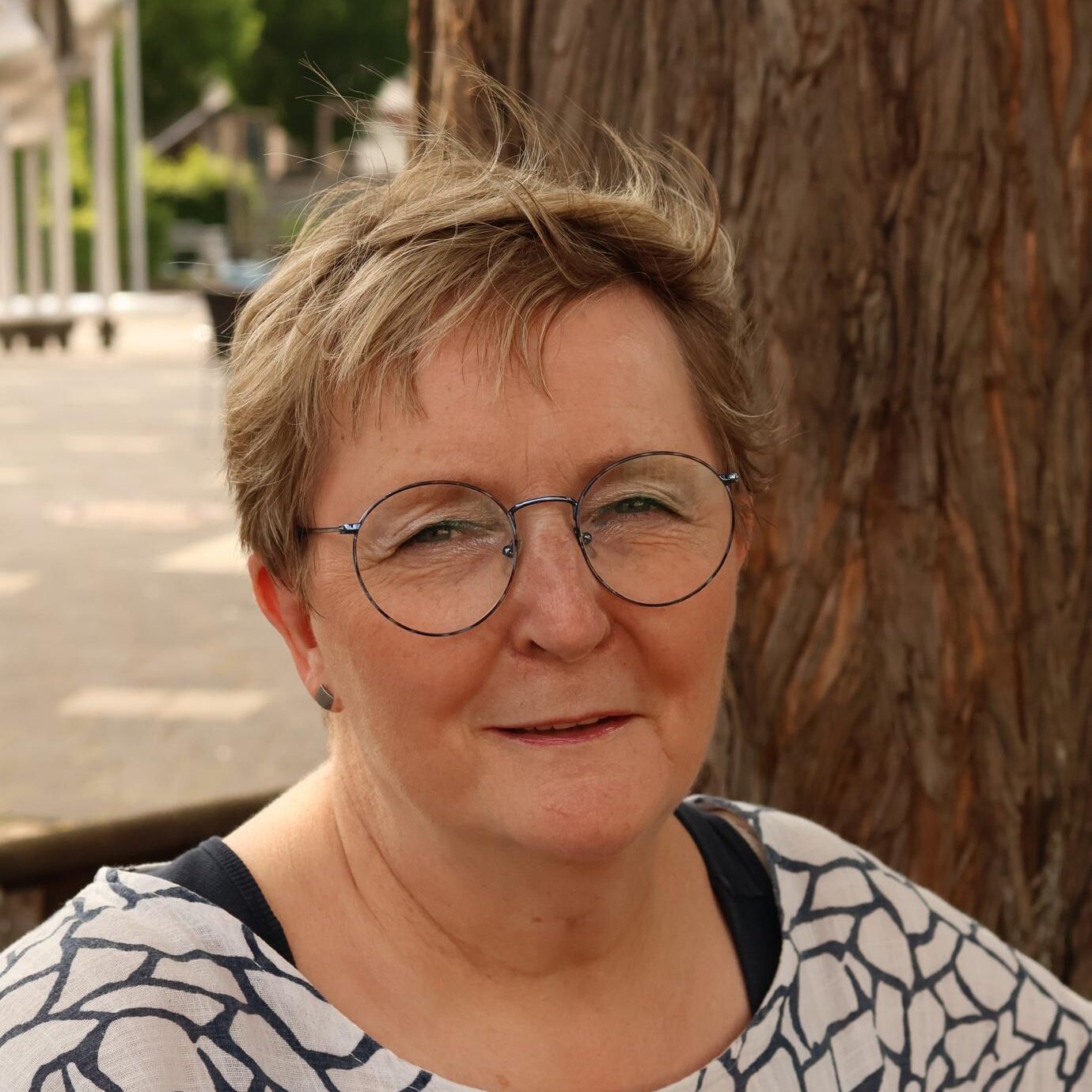 Susanne Petersohn Porträt einer Frau mit kurzem, blonden Haar und runden Brillen, die lächelt. Sie trägt ein weißes Oberteil mit schwarzem Muster und sitzt vor einem Baum.