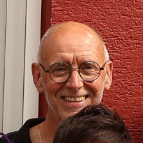 Lächelnder älterer Mann mit Brille, steht vor einer roten Wand. Er trägt ein schwarzes Polo-Shirt und hat eine freundliche Ausstrahlung.