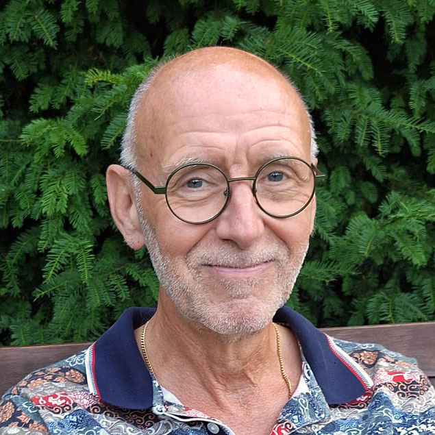 Frank Everts Ein älterer Mann mit Brille und einem freundlichen Lächeln sitzt vor einer grünen Hintergrundpflanze. Er trägt ein bunt gemustertes Hemd und einen goldenen Halsreifen.