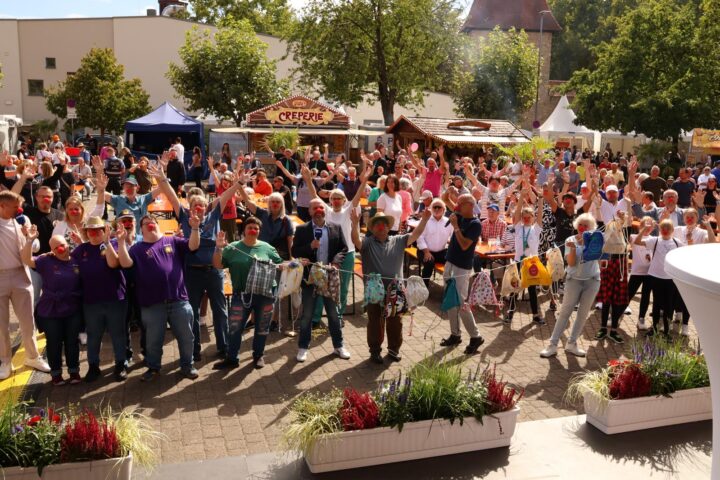 Gruppenszene bei einer Festveranstaltung mit Teilnehmern in bunten T-Shirts und roten Nasen, die begeistert die Arme heben. Im Hintergrund sind Essensstände und Zuschauer zu sehen.