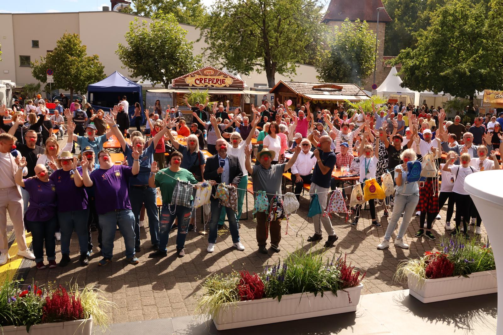 Gruppenszene bei einer Festveranstaltung mit Teilnehmern in bunten T-Shirts und roten Nasen, die begeistert die Arme heben. Im Hintergrund sind Essensstände und Zuschauer zu sehen.