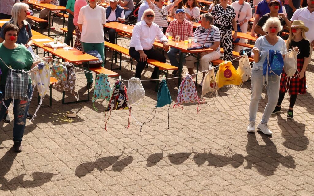 Menschen mit roten Clownsnasen präsentieren bunte Stoffbeutel, die an einer Schnur hängen, während Zuschauer an Tischreihen im Hintergrund sitzen.