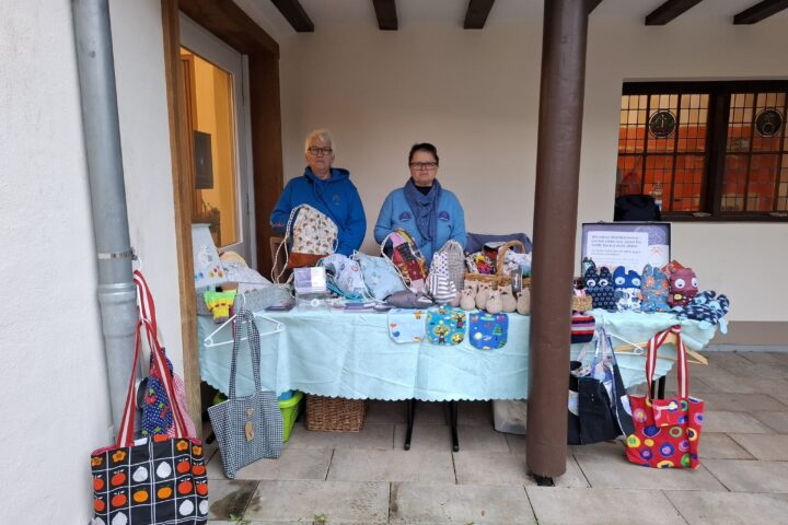 Stand mit handgefertigten Taschen und Accessoires, betrieben von zwei Frauen in blauen Jacken. Im Hintergrund sind verschiedene bunte Taschen und Körbe auf einem Tisch ausgestellt.