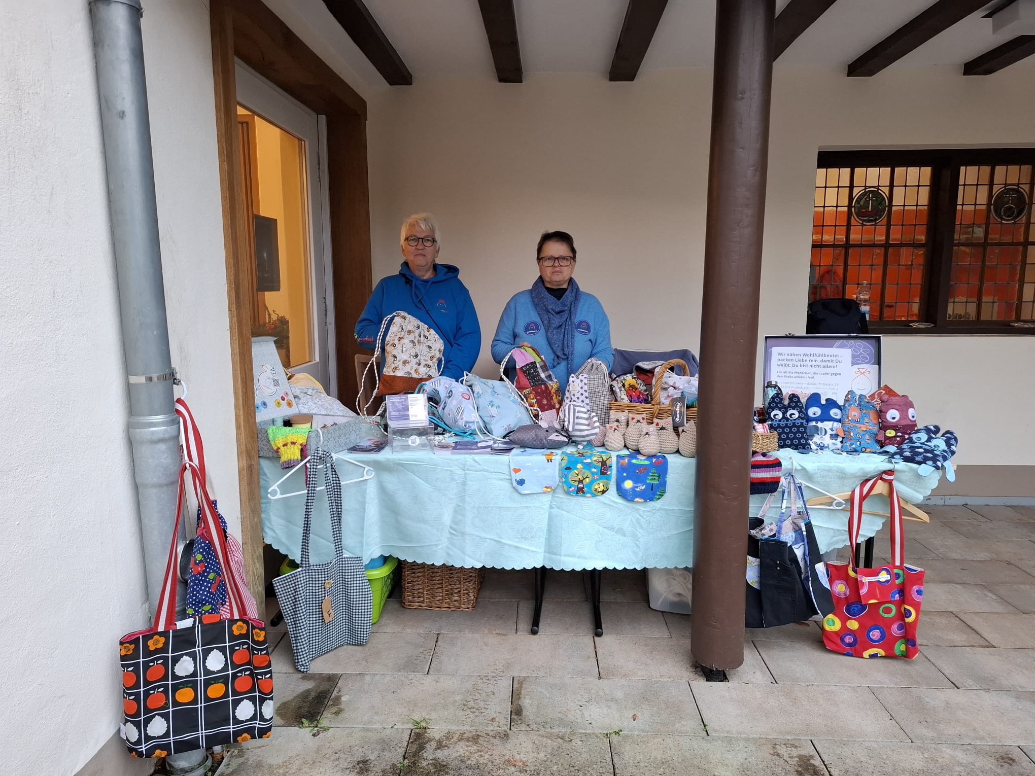 Stand mit handgefertigten Taschen und Accessoires, betrieben von zwei Frauen in blauen Jacken. Im Hintergrund sind verschiedene bunte Taschen und Körbe auf einem Tisch ausgestellt.