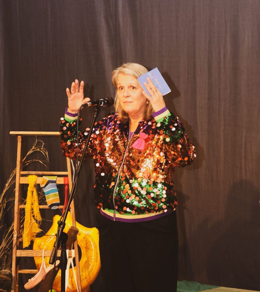 Frau mit blonder Haarfarbe und buntem Pailletten-Jackett, die am Mikrofon steht und ein Buch in der Hand hält. Hintergrund mit Holzregal und bunten Requisiten.