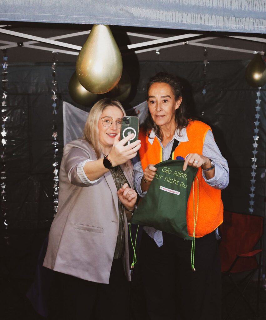 Zwei Frauen machen ein Selfie in einem festlich dekorierten Zelt. Eine trägt eine orange Weste und hält einen grünen Beutel mit dem Aufdruck "Gib alles zur nicht au". Die Szene vermittelt eine freundliche, gesellige Stimmung.