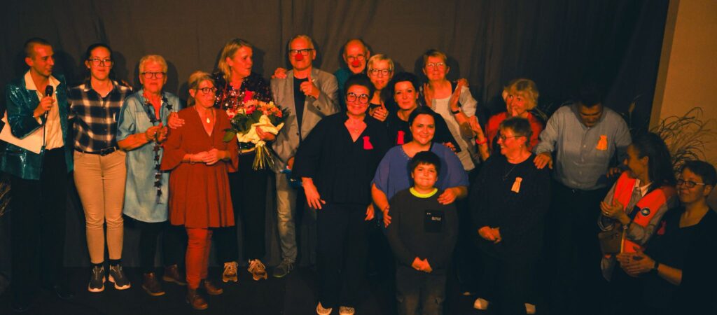 Gruppenszene mit mehreren Personen auf einer Bühne, einige tragen rote und orangefarbene Bänder. Im Vordergrund steht ein Junge, umgeben von Erwachsenen, die Blumen und Geschenke halten. Die Atmosphäre wirkt festlich und fröhlich.