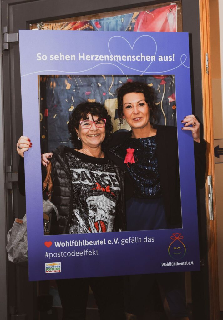 Zwei Frauen posieren in einem bunten Rahmen mit dem Text "So sehen Herzensmenschen aus!" Die Frau links trägt ein glitzerndes Oberteil mit einem Cartoon-Motiv und eine Brille, während die andere in einem eleganten Outfit mit einem roten Accessoire zu sehen ist. Der Kontext deutet auf eine Veranstaltung von Wohlfühlbeutel e.V. hin.